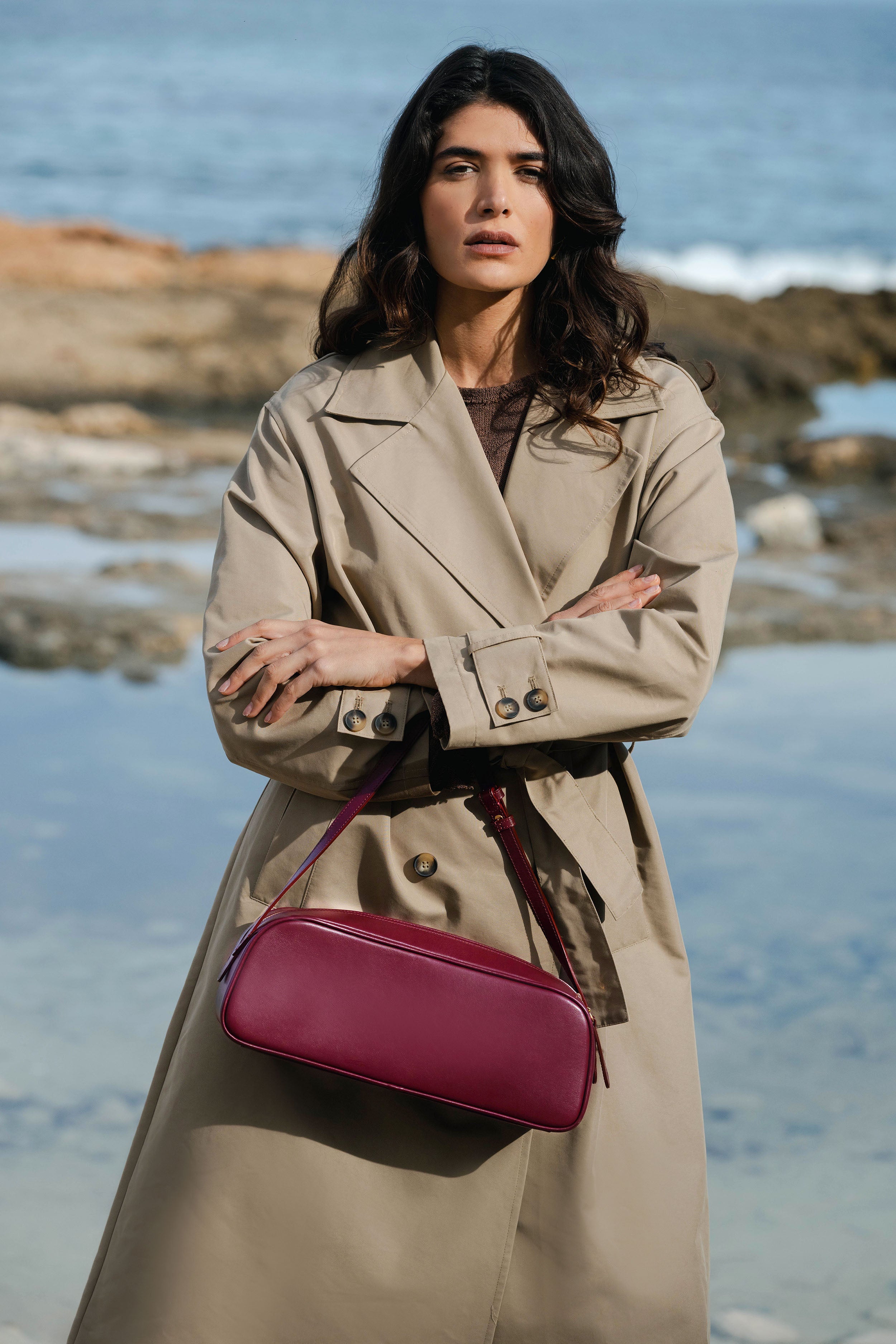 Woman in a beige trench coat with a red handbag standing on a rocky beach.