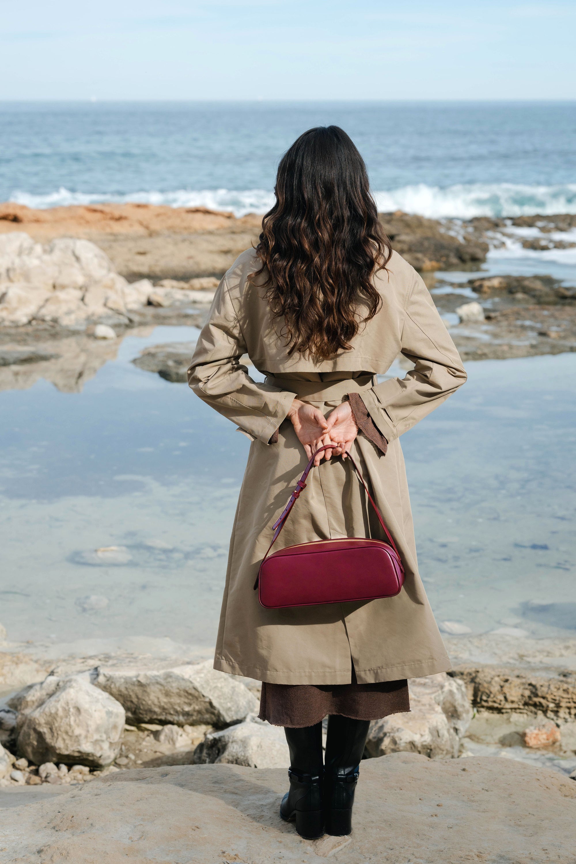 Woman in a trench coat holding a red handbag by the ocean