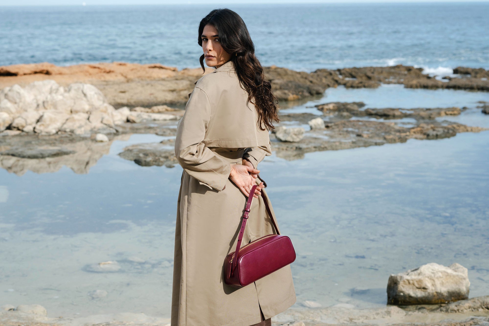 Woman in a trench coat holding a red handbag by the sea.